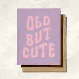 The “Old But Cute” birthday card by Daydream Prints features retro pink text on lavender, shown with a brown envelope against a light background.