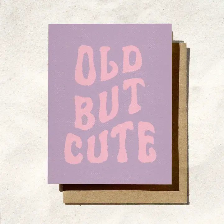 The “Old But Cute” birthday card by Daydream Prints features retro pink text on lavender, shown with a brown envelope against a light background.