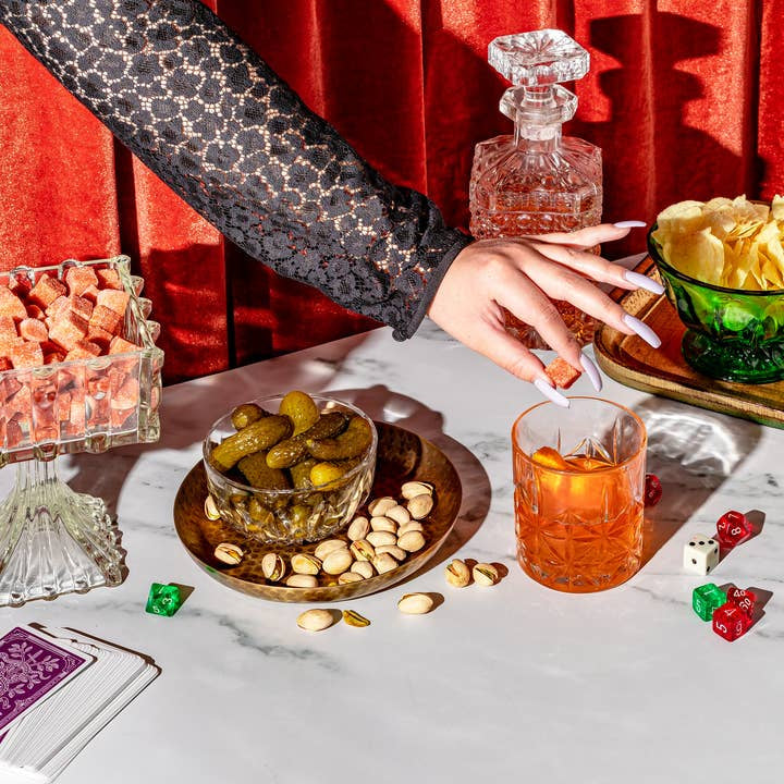 A hand reaches for a glass of Old Fashioned made with Yes Cocktail Co.'s Old Fashioned Cocktail Cubes on a table with snacks, dice, cards, and a decanter, set against a red velvet curtain.