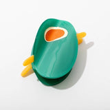 A toy avocado with removable pit and yellow handles sits on a white surface next to the Olive Claw Clip by Larissa Loden, an elegant handmade hair accessory crafted from cellulose acetate.