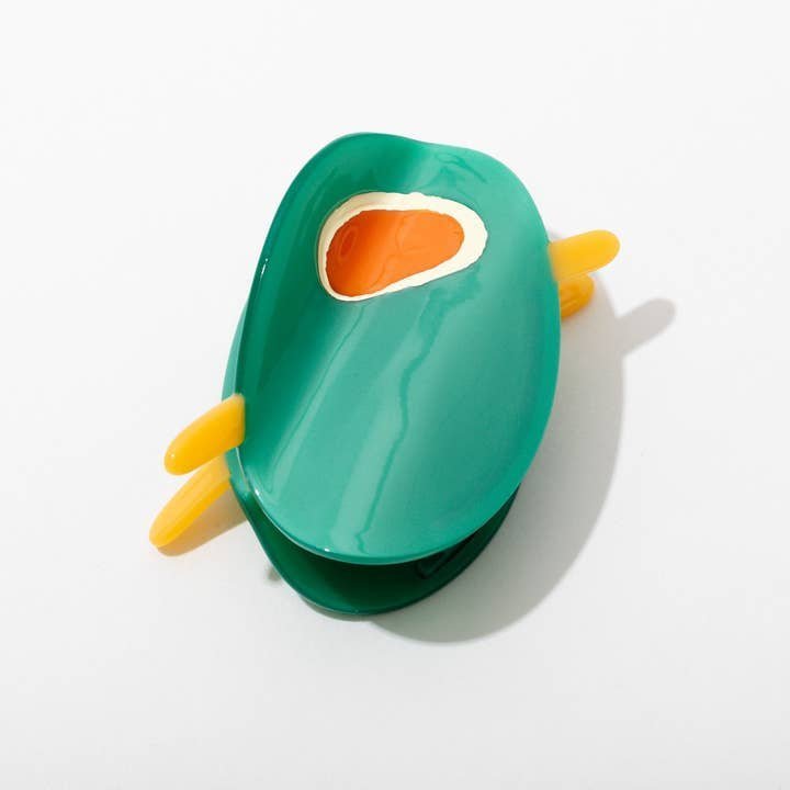 A toy avocado with removable pit and yellow handles sits on a white surface next to the Olive Claw Clip by Larissa Loden, an elegant handmade hair accessory crafted from cellulose acetate.