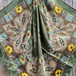 The Olive Tone Paisley Wild Rag by Rodeo Drive is a square silky polyester scarf, featuring paisley and floral prints in olive green, brown, yellow, and blue—styled for western wear and shown gathered at the center on wood.