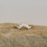 JaxKelly's Opal Gold Ring features five marquise-shaped white stones set in a fan pattern and is displayed on a rough beige stone surface.
