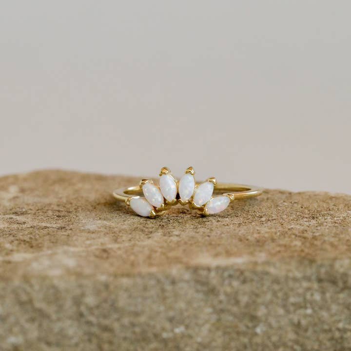 JaxKelly's Opal Gold Ring features five marquise-shaped white stones set in a fan pattern and is displayed on a rough beige stone surface.