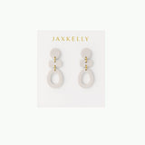 The Organic Three Tier Clay Earring in Oat Milk by JaxKelly features geometric, off-white polymer clay tiers with gold connectors—stylish, lightweight earrings on a branded card, ideal for everyday wear.