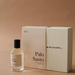 A bottle of Palo Santo Eau De Parfum by dilo, a woody scent with sandalwood notes, rests beside its beige box adorned with the phrase "to the elsewhere..." on the lid.