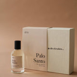 A bottle of Palo Santo Eau De Parfum by dilo, a woody scent with sandalwood notes, rests beside its beige box adorned with the phrase "to the elsewhere..." on the lid.