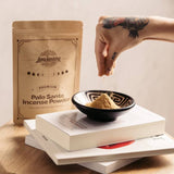 A hand sprinkles Luna Sundara's Palo Santo Incense Powder into a black and white bowl, with the branded pouch, books, and a wooden surface in the background.