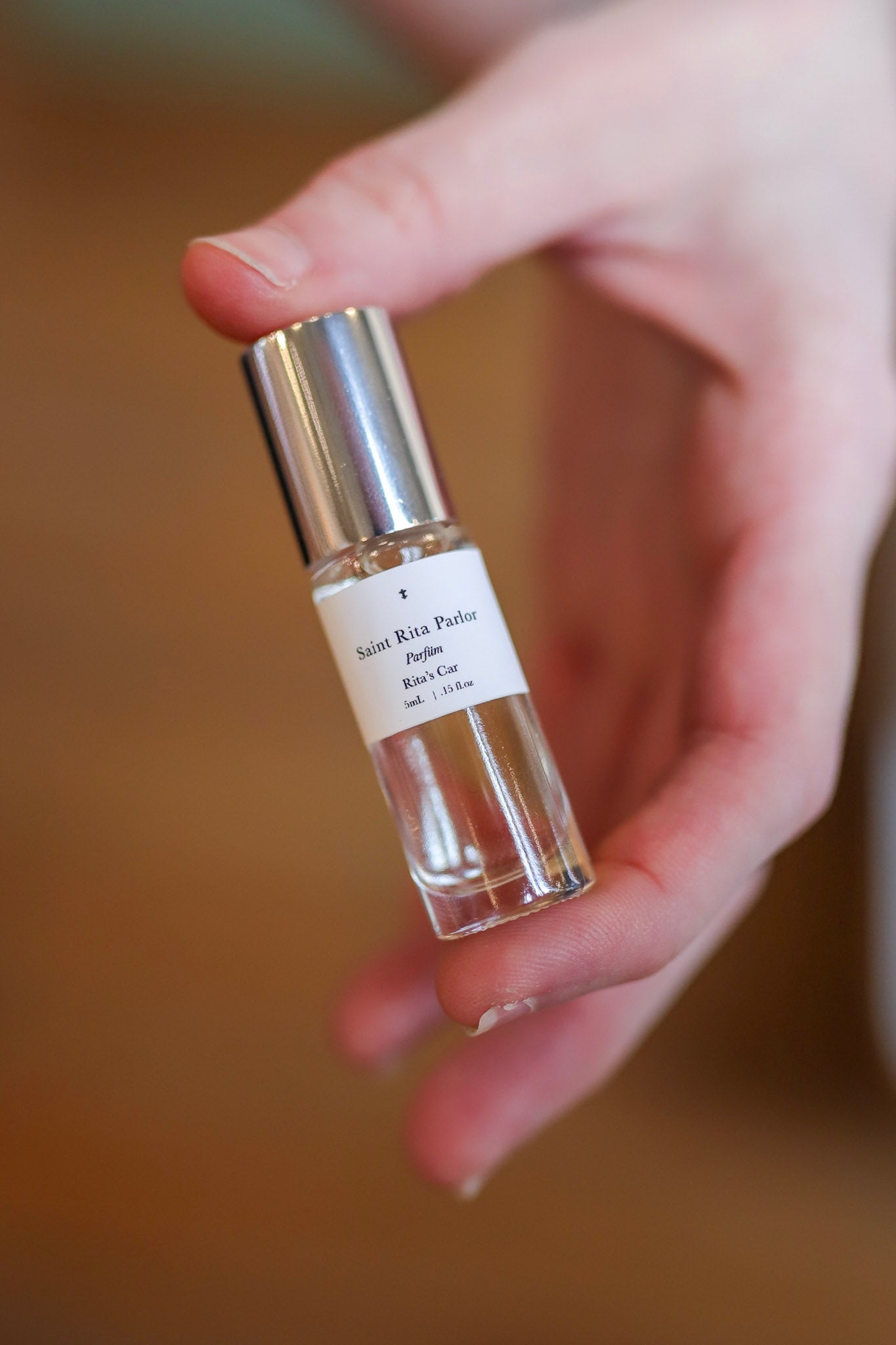 A hand holds a small glass bottle of "Parfum | Rita’s Car" by Saint Rita Parlor, with a silver cap and white label, containing a unisex fragrance made from organic essential oils.