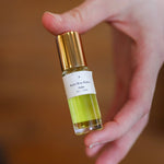 A hand holds a small glass bottle of Saint Rita Parlor’s Signature Parfum, featuring a gold cap and light green liquid—a unique unisex fragrance crafted from organic essential oils.