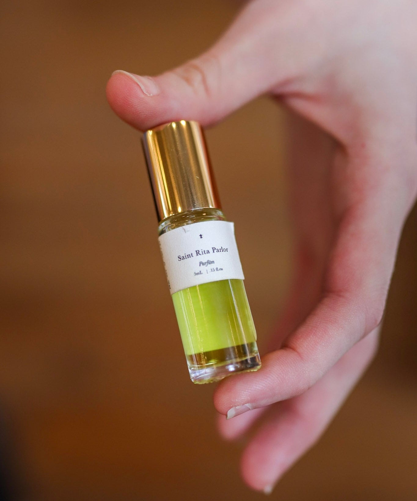 A hand holds a small glass vial of Saint Rita Parlor’s Parfum | Signature, featuring a gold cap and yellow-green liquid inside.