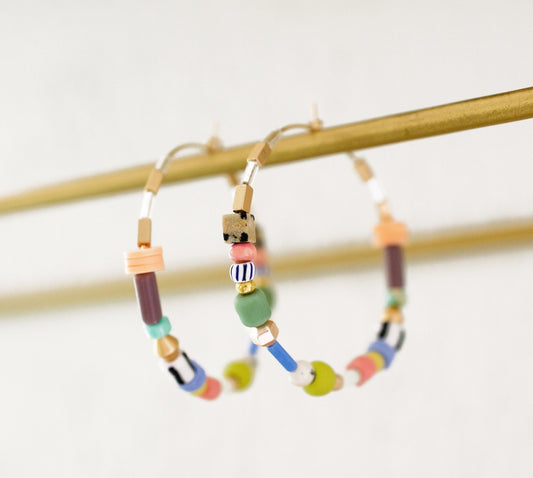 The Pastel Beaded Hoops by Jill Makes feature large 45mm hoops adorned with multicolored beads in assorted shapes and sizes, all hanging from a horizontal gold rod.