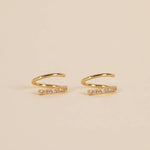 The Pave Spiral - Gold Earrings by JaxKelly are a pair of hypoallergenic gold ear cuffs with a curved design and a row of clear stones on one side, shown on a beige background.