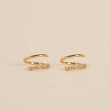 The Pave Spiral - Gold Earrings by JaxKelly are a pair of hypoallergenic gold ear cuffs with a curved design and a row of clear stones on one side, shown on a beige background.