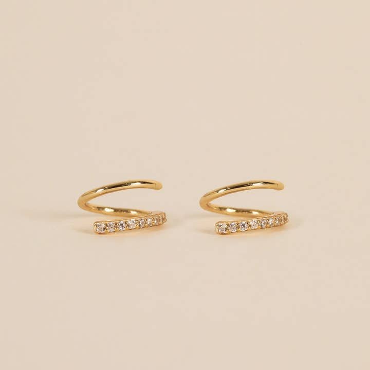The Pave Spiral - Gold Earrings by JaxKelly are a pair of hypoallergenic gold ear cuffs with a curved design and a row of clear stones on one side, shown on a beige background.