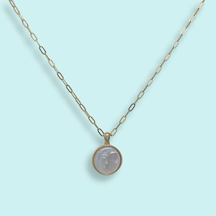 The Pearl Man-in-the-Moon Necklace by Ornamental Things features a gold chain and a round mother-of-pearl pendant with an embossed face design, shown on a light blue background.
