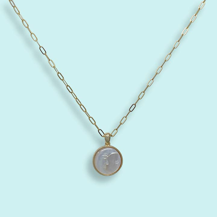 The Pearl Man-in-the-Moon Necklace by Ornamental Things is a gold chain necklace with a round mother-of-pearl pendant engraved with a face, shown on a light blue background.