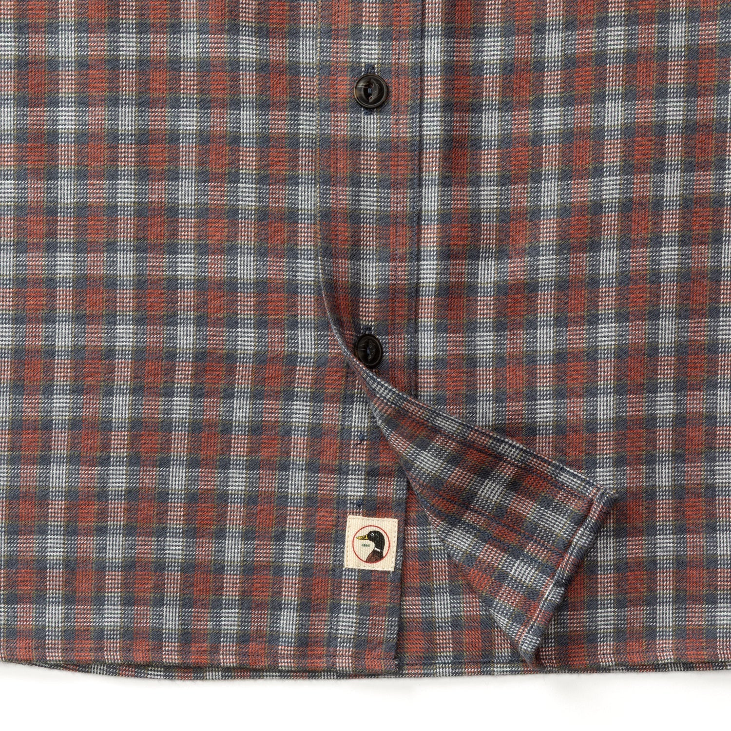 Close-up of the Duck Head Performance Flannel Sport Shirt, a brown, blue, and gray plaid moisture-wicking shirt with black buttons and a logo tag on the hem—ideal for outdoor hiking.