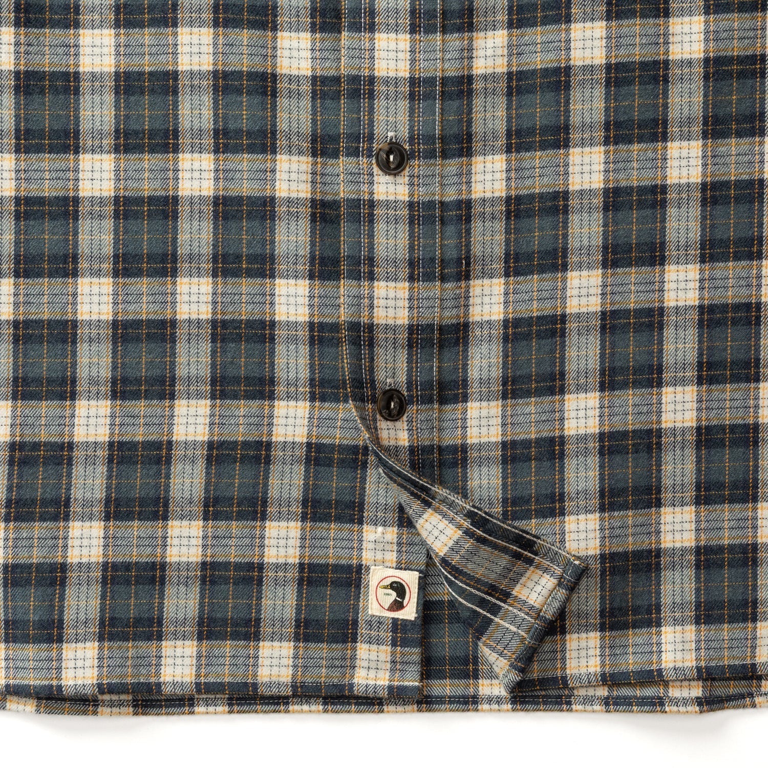 Close-up of the Duck Head Performance Flannel Sport Shirt in blue, beige, and white with black buttons and a small face icon tag near the hem.