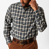 A man wears the Duck Head Performance Flannel Sport Shirt, a blue and gray plaid wool-polyester button-up with chest pockets, paired with brown pants, standing against a plain white background.