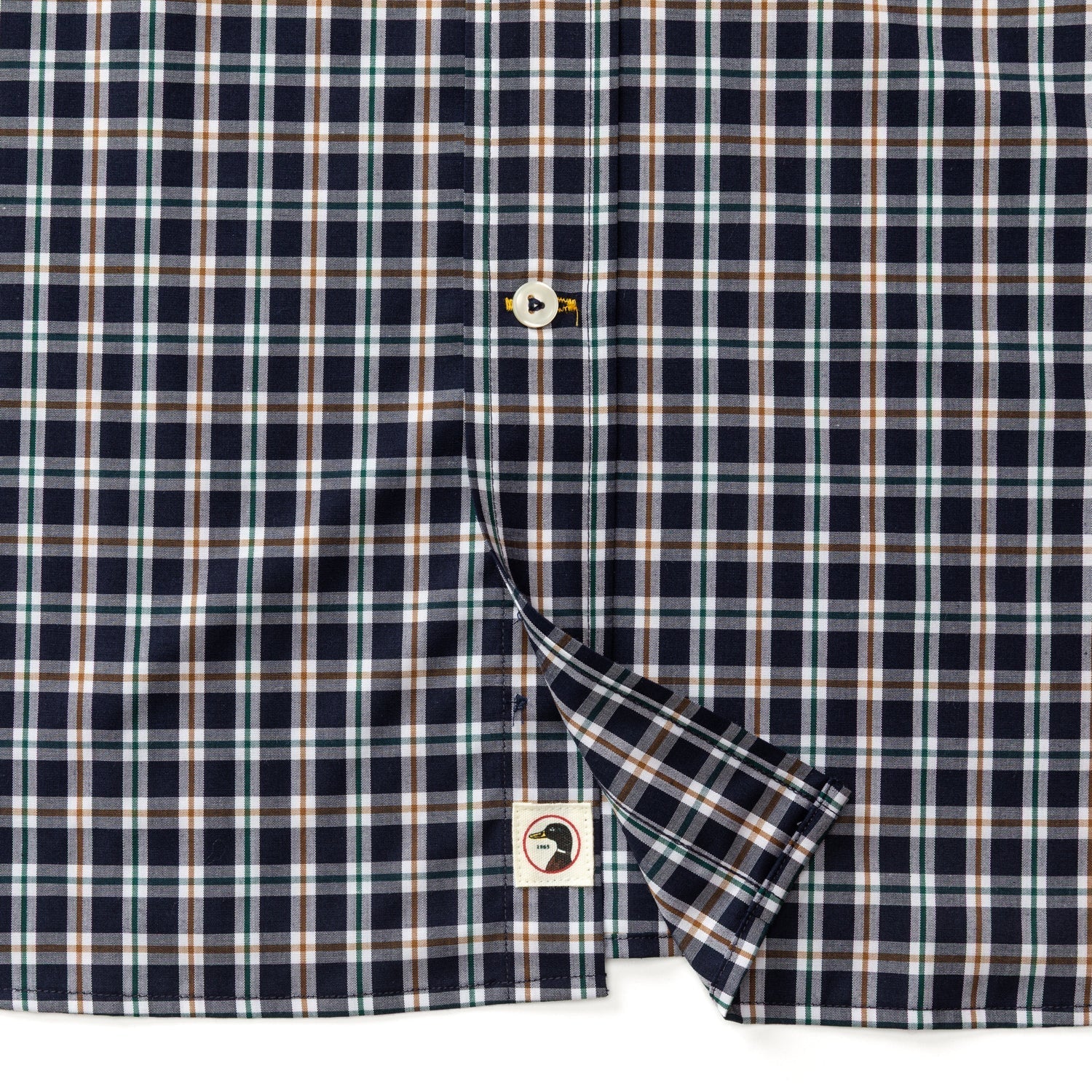 Close-up of the Duck Head Performance Poplin Sport Shirt, a navy, brown, and white plaid button-down with visible buttons and a small fabric tag on the hem.