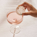 A hand places a round cocktail ice sphere, made with the Petal Ice Mold by W&P, into a glass of pink liquid on a white surface.