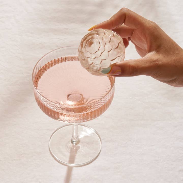 A hand holds a decorative round ice sphere made with the Petal Ice Mold by W&P above a glass of pink liquid on a white surface.
