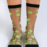 Feet in Tailored Union’s Petal Sheer Ankle Sock—sheer black socks with neon green floral patterns and solid, reinforced toes, heels, and cuffs for durability.