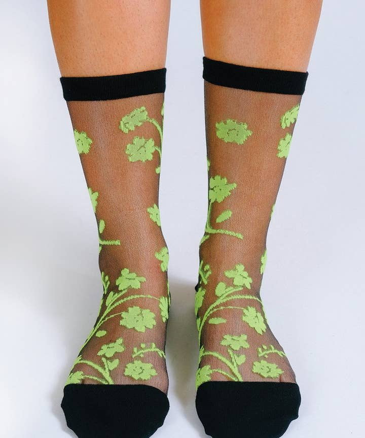 Feet in Tailored Union’s Petal Sheer Ankle Sock—sheer black socks with neon green floral patterns and solid, reinforced toes, heels, and cuffs for durability.