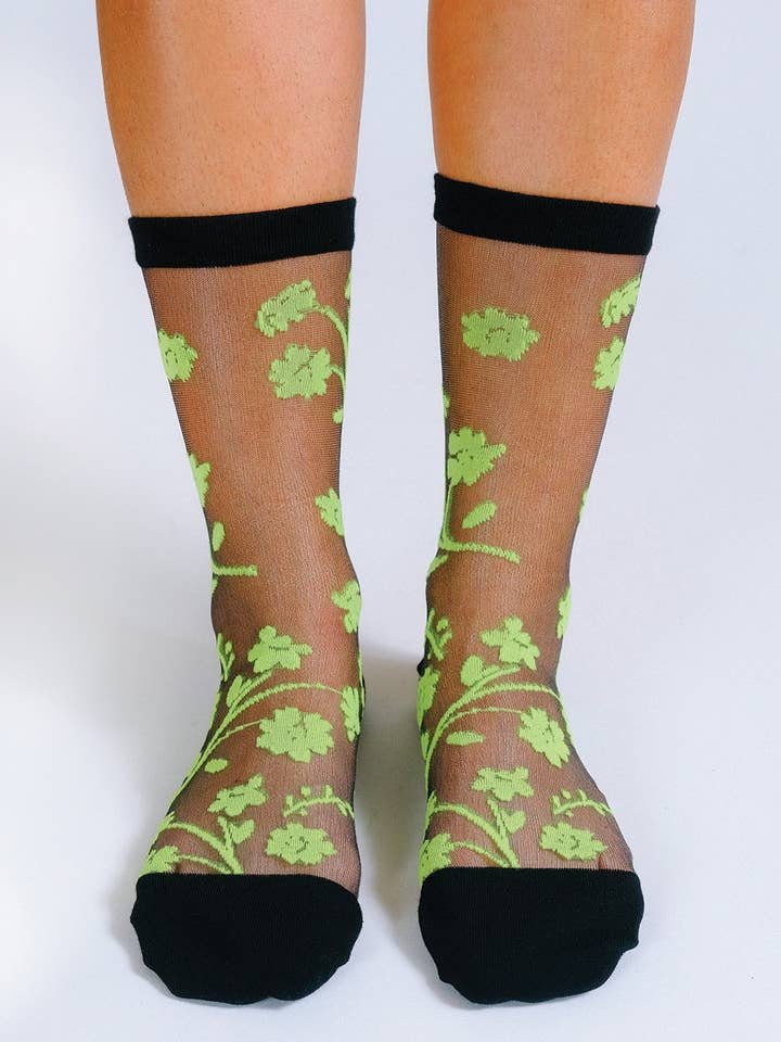 Feet in Tailored Union’s Petal Sheer Ankle Sock—sheer black socks with neon green floral patterns and solid, reinforced toes, heels, and cuffs for durability.
