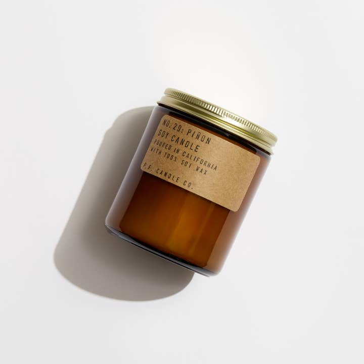 A brown amber jar candle with a gold metal lid and kraft label reading "Piñon | P.F Candle" from P.F Candle, featuring piñon, cedar, and vanilla notes, is shown on a white background.