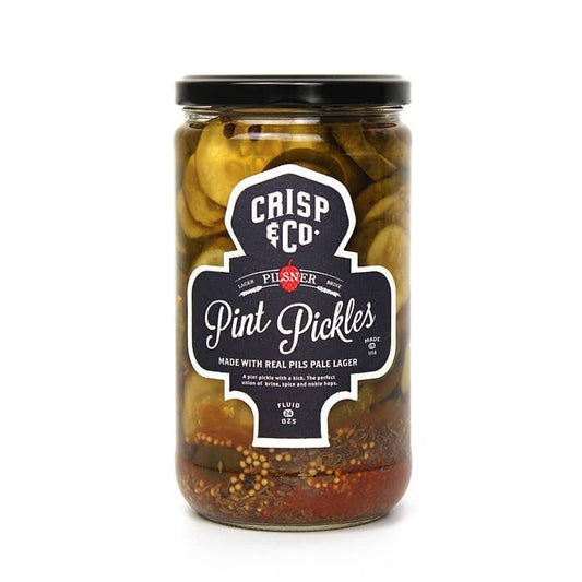 A glass jar of Crisp & Co. Pint Pickles features sliced pickles, dill seed, heirloom garlic, and spices in pilsner pale lager. The black label displays the Crisp & Co. brand name, product name, and ingredients.