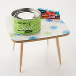 A can of Potato Salad Salt by Beautiful Briny Sea sits beside two boxes of aluminum foil on a small, square table with a white and blue patterned surface and wooden legs.