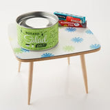 A jar of Potato Salad Salt by Beautiful Briny Sea sits on a small, square table with a blue and green pattern and wooden legs—perfect for seasoning grilled veggies or creating flavorful dishes.