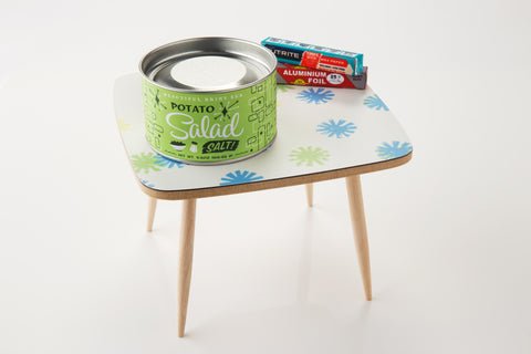 A jar of Potato Salad Salt by Beautiful Briny Sea sits on a small, square table with a blue and green pattern and wooden legs—perfect for seasoning grilled veggies or creating flavorful dishes.