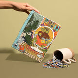 A hand holds a colorful Rainy Day 1,000 Pieces puzzle box by Birdie Puzzles, featuring an illustrated design, next to a tipped-over cup spilling recycled puzzle pieces onto a flat surface.