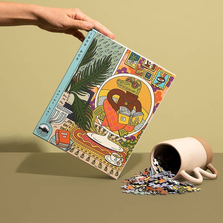A hand holds a colorful Rainy Day 1,000 Pieces puzzle box by Birdie Puzzles, featuring an illustrated design, next to a tipped-over cup spilling recycled puzzle pieces onto a flat surface.