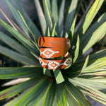 The Rambler by Zilker Belts is a hand-stitched Argentine leather belt featuring white and orange geometric designs and "Zilker" embossed, shown on vibrant green leaves.