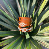 The Rambler by Zilker Belts is a hand-stitched Argentine leather belt featuring white and orange geometric designs and "Zilker" embossed, shown on vibrant green leaves.