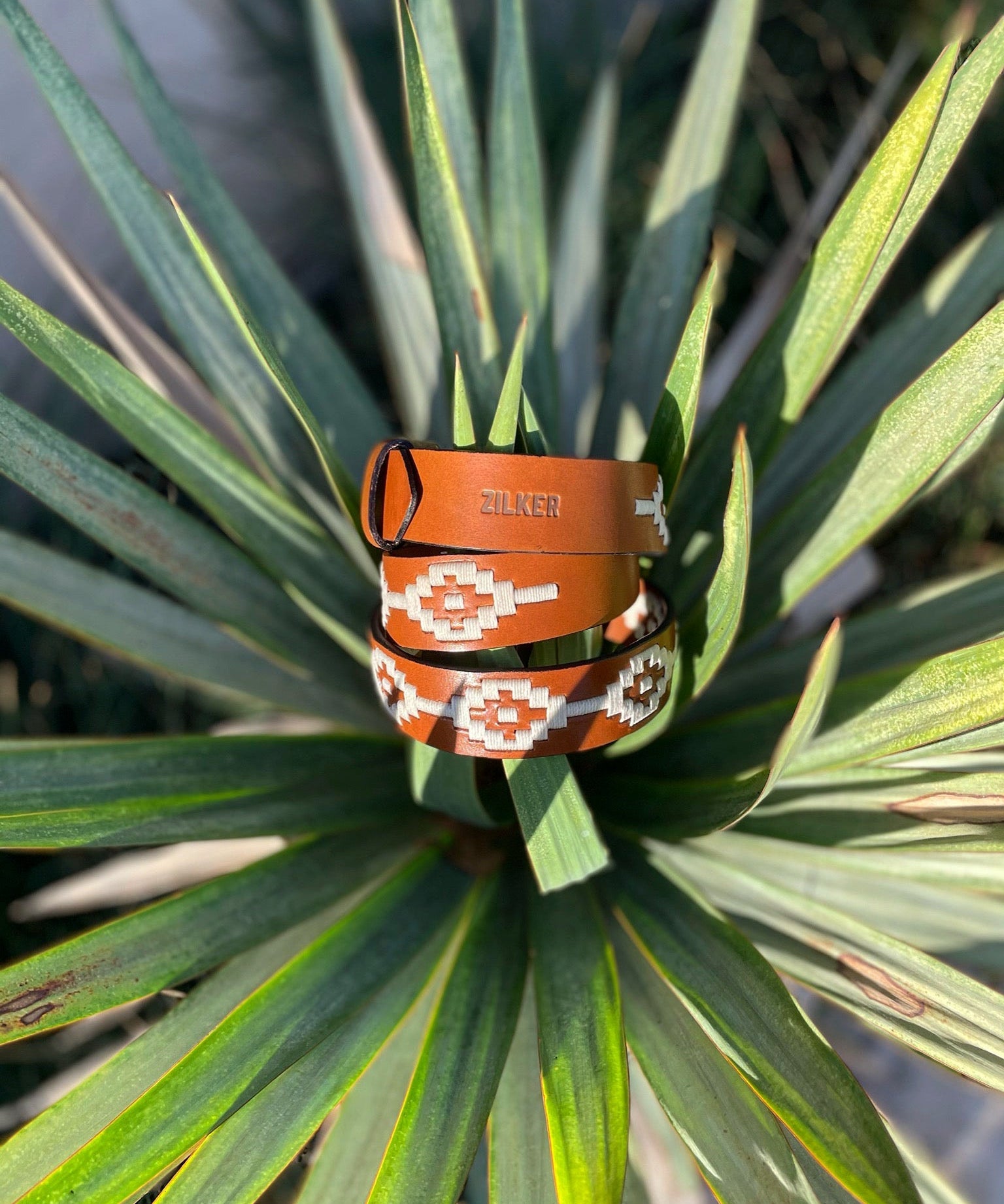 The Rambler by Zilker Belts is a hand-stitched Argentine leather belt featuring white and orange geometric designs and "Zilker" embossed, shown on vibrant green leaves.