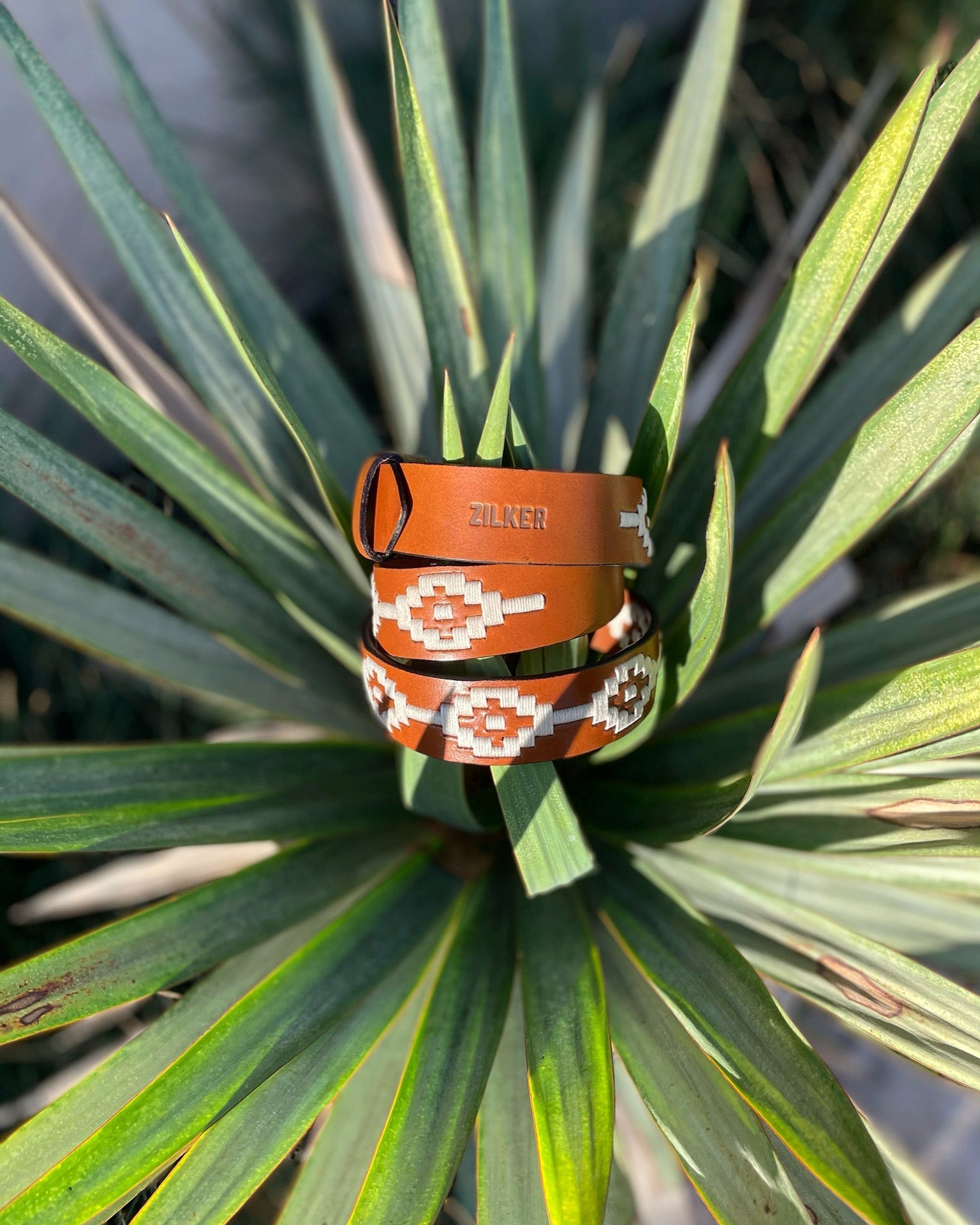 The Rambler by Zilker Belts is a hand-stitched Argentine leather belt featuring white and orange geometric designs and "Zilker" embossed, shown on vibrant green leaves.