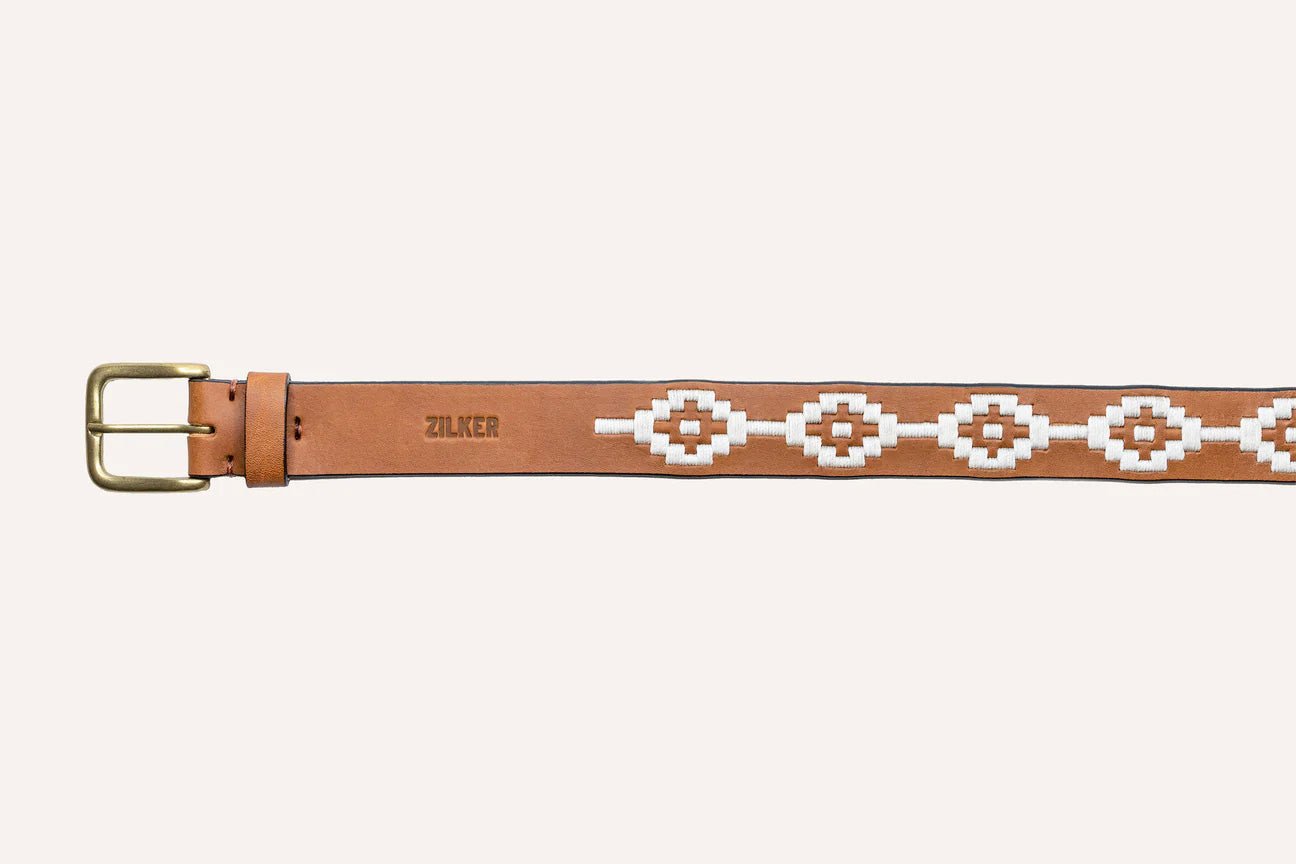 The Rambler by Zilker Belts is a light brown Argentine leather belt with a brass buckle, white geometric bead pattern, and "ZULKER" embossed near the buckle—distinctively hand-stitched.