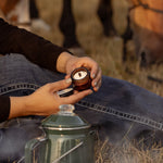 Seated on dry grass, a person holds a small RANCH ROAST Coffee & Vanilla Infused Tallow Eye Cream by Cowboy & Co., with a green metal coffee pot in the foreground.