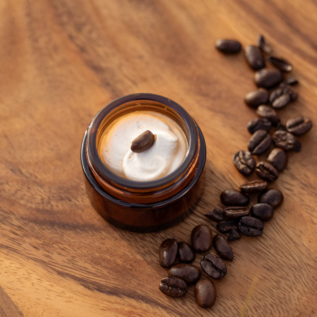 The RANCH ROAST Coffee & Vanilla Infused Tallow Eye Cream by Cowboy & Co. sits in a small brown jar on a wooden surface, with coffee beans scattered nearby and a coffee bean on the lid.