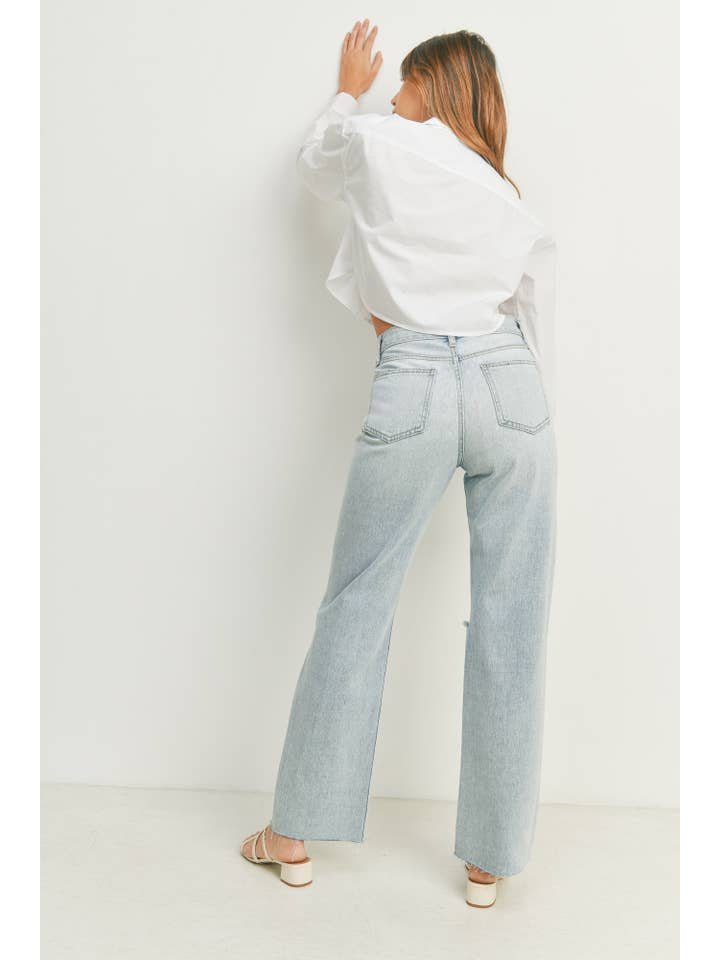 A woman in a white shirt and JBD Ribcage Longer Length Straight jeans in light denim (32-inch inseam) stands facing a white wall with her back to the camera, one hand raised.