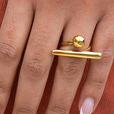 A hand wears the Peter and June Balance Ring, an adjustable, waterproof 18k gold-plated stainless steel piece with round and horizontal bar design, resting on rust-colored fabric—everyday elegance by Peter and June.