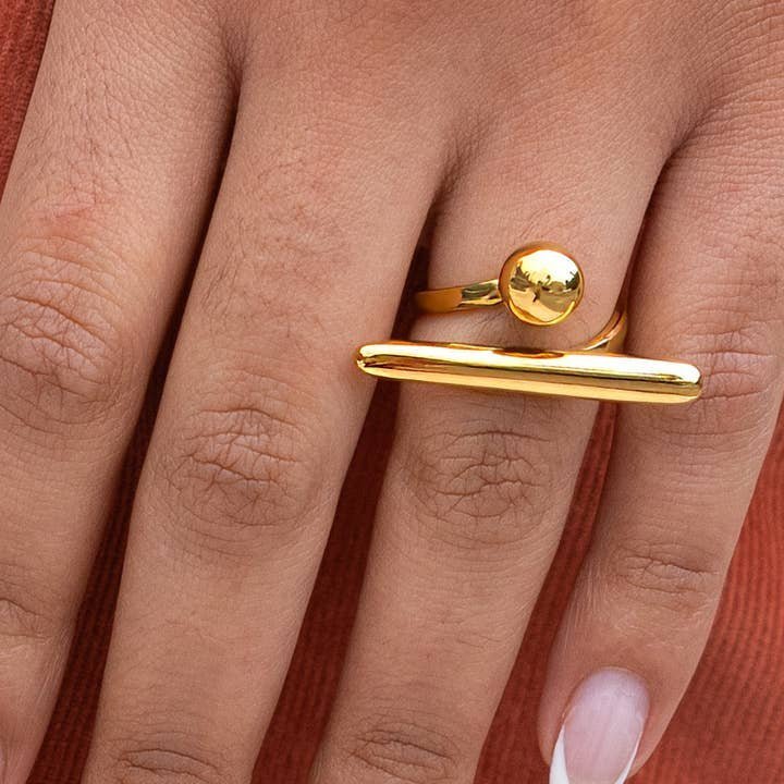 A hand wears the Peter and June Balance Ring, an adjustable, waterproof 18k gold-plated stainless steel piece with round and horizontal bar design, resting on rust-colored fabric—everyday elegance by Peter and June.