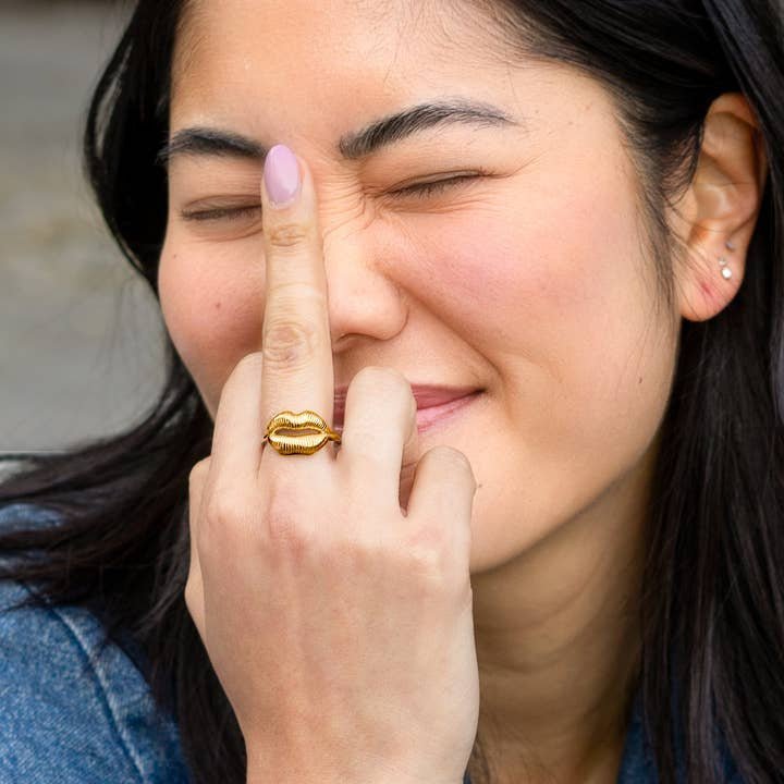 A person with long dark hair, eyes closed and smiling, shows their middle finger adorned with Peter and June’s Ring | Gold Lips—an ethically sourced, 18k gold-plated adjustable ring shaped like lips.