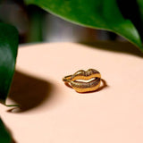 The Ring | Gold Lips by Peter and June, an adjustable 18k gold plated ring shaped like lips, is displayed on a beige surface with green leaves nearby.