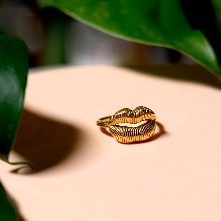 The Ring | Gold Lips by Peter and June, an adjustable 18k gold plated ring shaped like lips, is displayed on a beige surface with green leaves nearby.